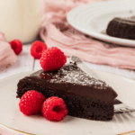 piece of flourless chocolate cake on a white plate topped with powdered sugar and raspberries
