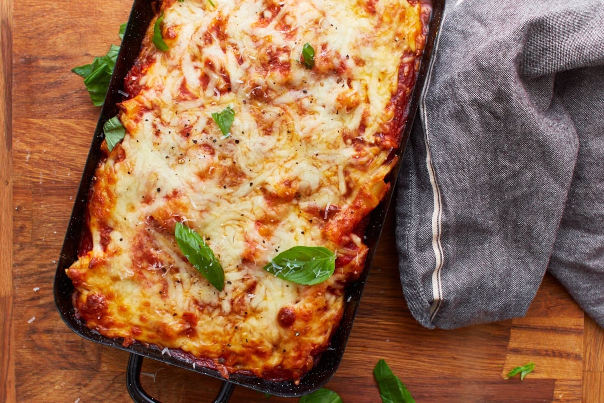 overhead view of baked gluten-free baked ziti in black baking dish on wooden counter with gray rag next to it.