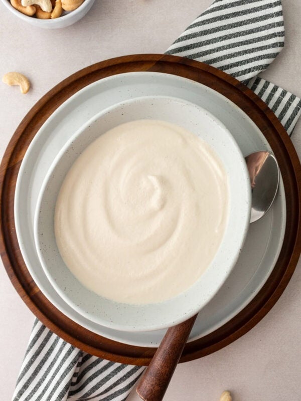 A bowl of cashew cream with a silky smooth surface, placed on a wooden tray and surrounded by cashew nuts and a striped napkin.