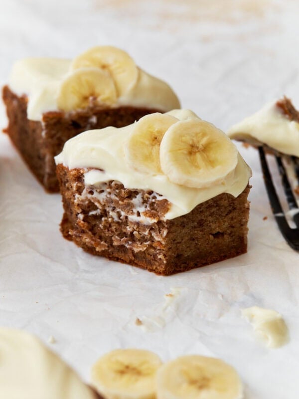 A close-up of a slice of gluten-free banana cake topped with creamy frosting and fresh banana slices, resting on parchment paper with a fork in the background