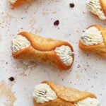 A close-up image of crispy, golden gluten-free cannoli shells filled with piped creamy ricotta filling, garnished with chocolate chips and dusted with cocoa powder on a light-textured surface.