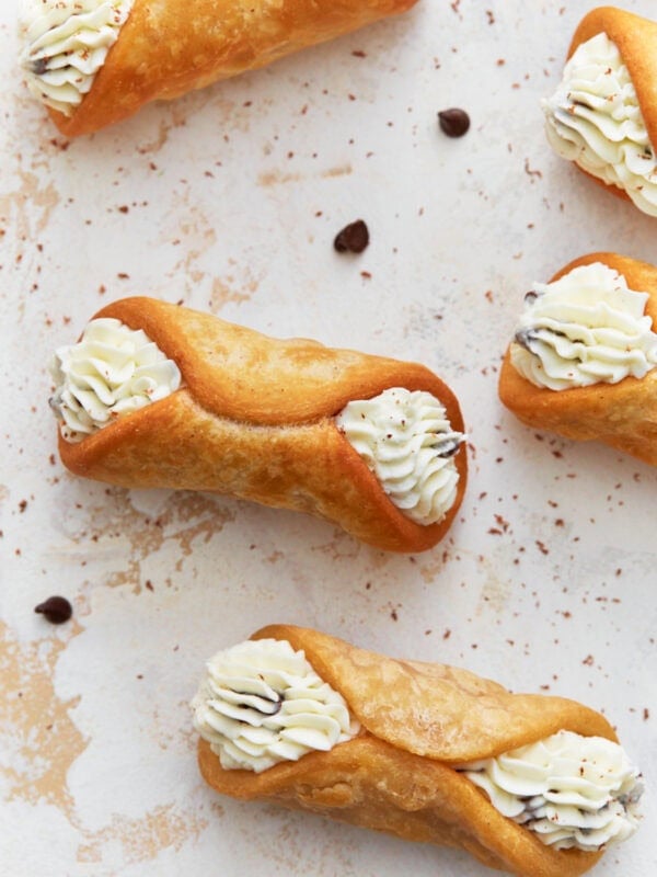 A close-up image of crispy, golden gluten-free cannoli shells filled with piped creamy ricotta filling, garnished with chocolate chips and dusted with cocoa powder on a light-textured surface.