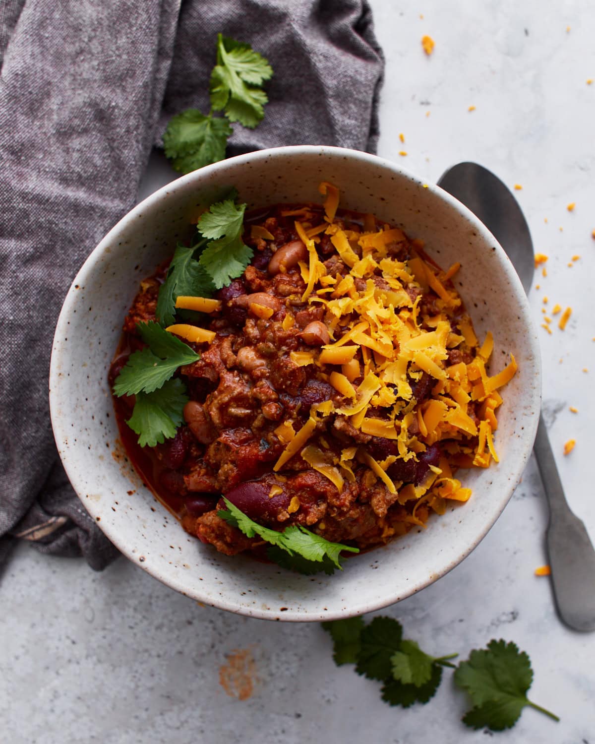 A white bowl filled with gluten-free chili, topped with parsley and shredded orange cheese. The bowl is resting on a dark grey towel with a spoon resting on the light counter beside it.