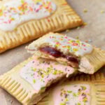Homemade gluten-free pop tarts with white icing and colorful sprinkles on top, filled with a vibrant red jam, displayed on a parchment-lined surface. One pop tart is halved to reveal the jam filling inside