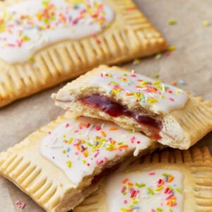 Homemade gluten-free pop tarts with white icing and colorful sprinkles on top, filled with a vibrant red jam, displayed on a parchment-lined surface. One pop tart is halved to reveal the jam filling inside