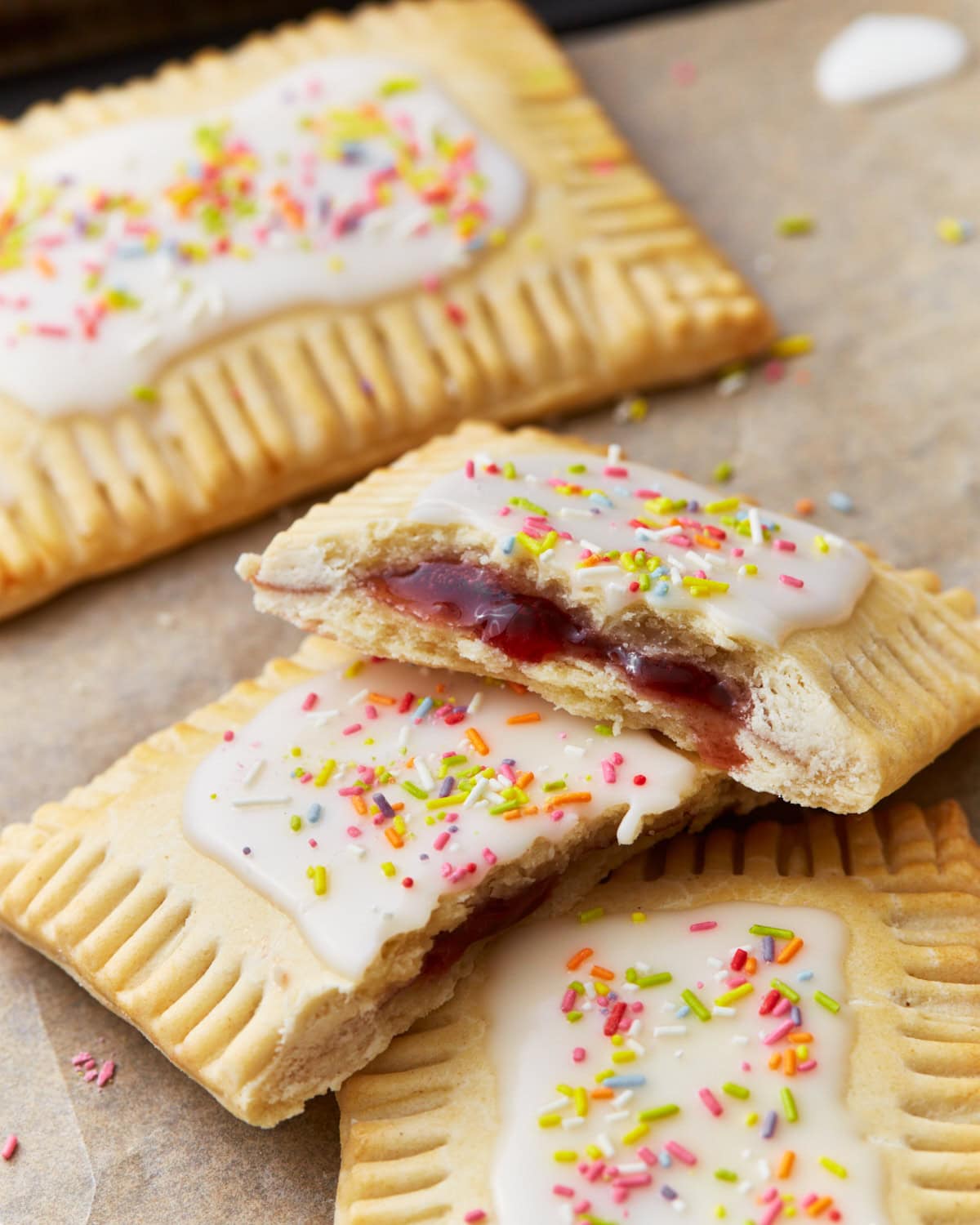Homemade gluten-free pop tarts with white icing and colorful sprinkles on top, filled with a vibrant red jam, displayed on a parchment-lined surface. One pop tart is halved to reveal the jam filling inside