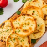 Golden brown arepas served on a wooden board, garnished with fresh chopped parsley. Fresh tomatoes and cilantro leaves are in the background, adding a vibrant touch.