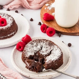 A gluten-free chocolate lava cake on a white plate, dusted with powdered sugar, surrounded by fresh raspberries and chocolate chips. A fork reveals the gooey chocolate center. A pink cloth and a bottle of milk in the background add a soft touch.