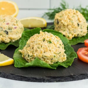 Close-up of vegan chickpea 'tuna' salad scoops served on fresh lettuce leaves, garnished with dill, lemon wedges, and tomato slices on a black slate platter.
