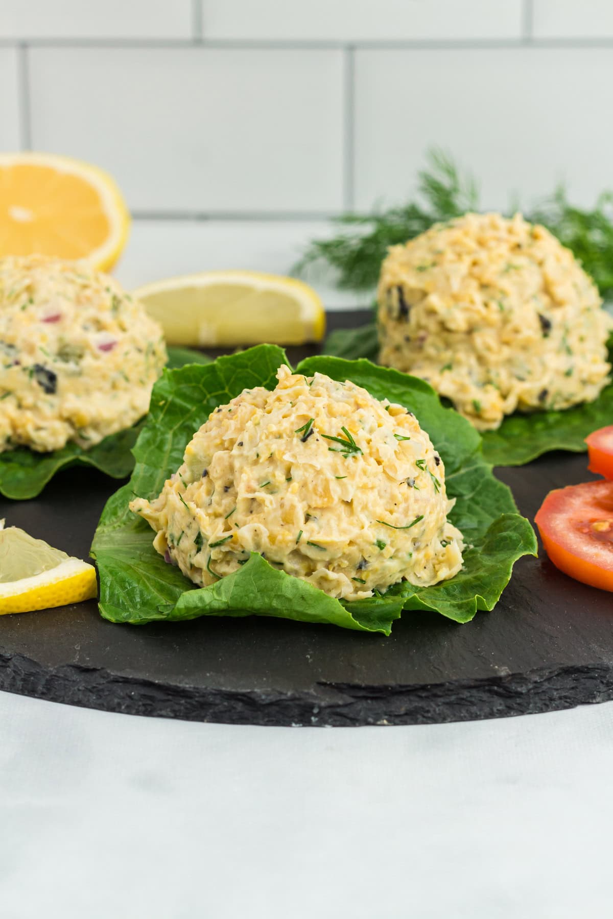 Close-up of vegan chickpea 'tuna' salad scoops served on fresh lettuce leaves, garnished with dill, lemon wedges, and tomato slices on a black slate platter.