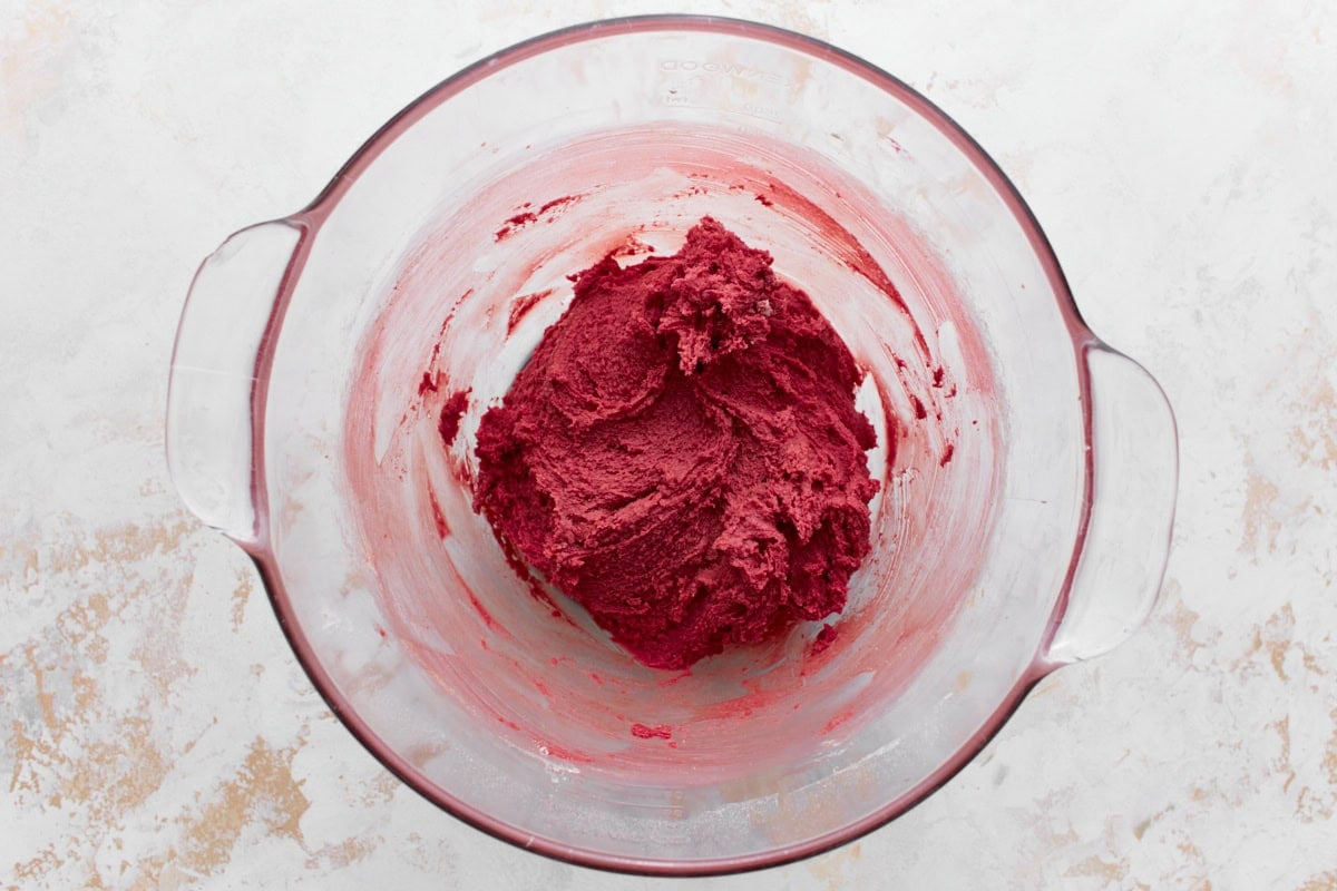 A glass mixing bowl containing vibrant red cake batter, thick and smooth in texture, sitting on a textured white and beige surface.






