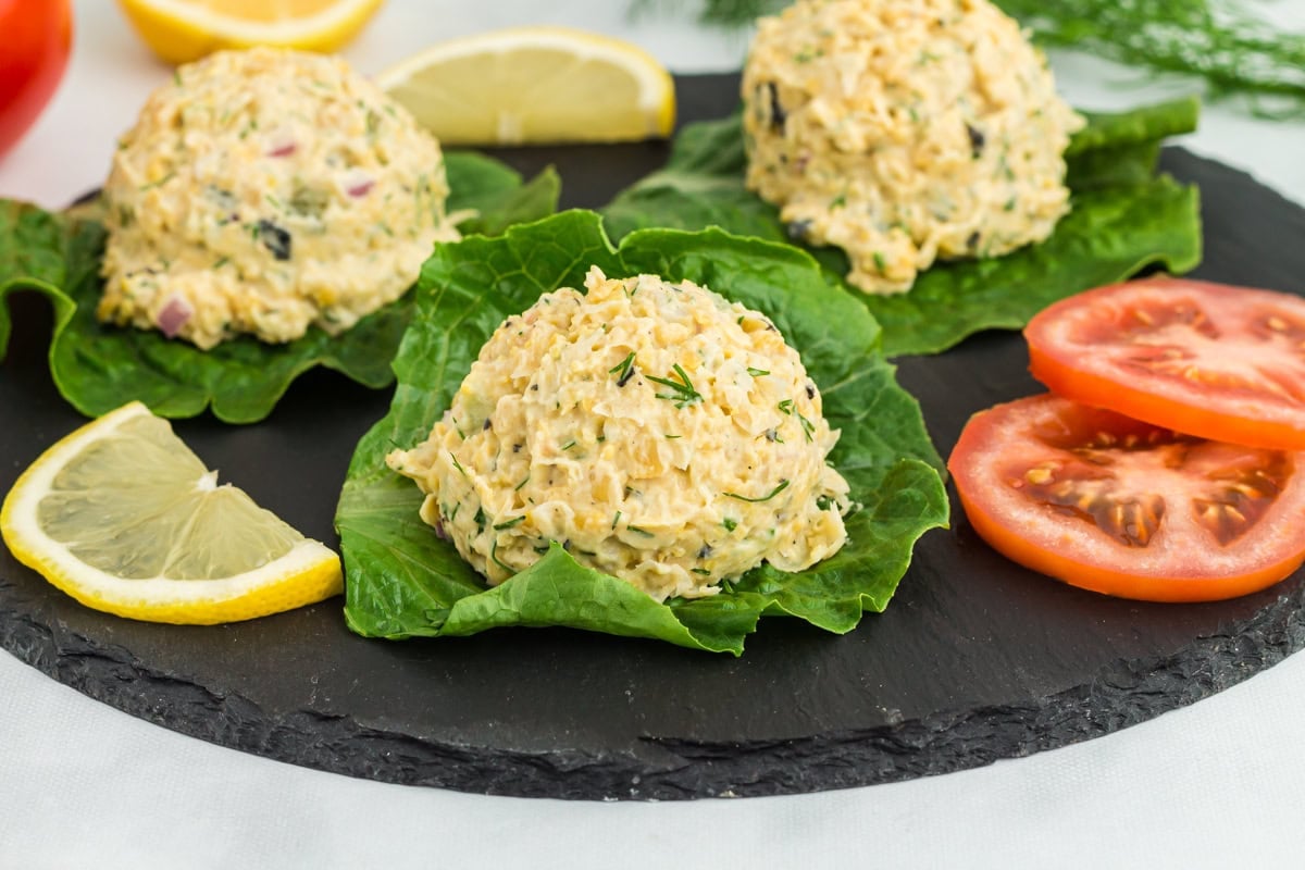 Vegan chickpea 'tuna' salad scoops served on fresh lettuce leaves, garnished with dill, lemon wedges, and tomato slices on a black slate platter.