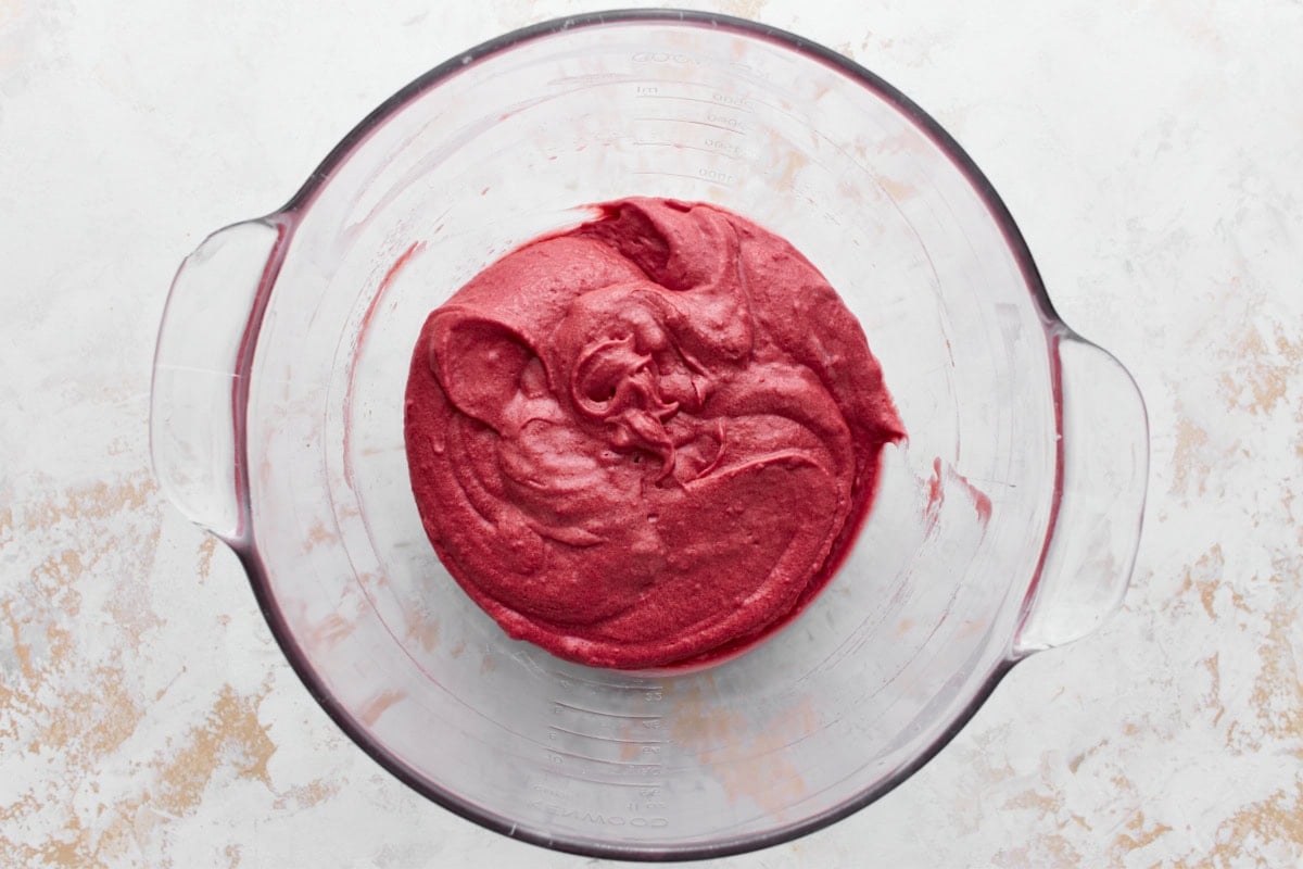 A glass mixing bowl containing smooth gluten-free red velvet cake batter, sitting on a textured white and beige surface.






