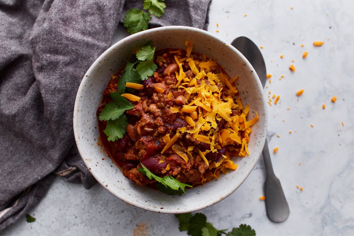 A white bowl filled with gluten-free chili, topped with parsley and shredded orange cheese. The bowl is resting on a dark grey towel with a spoon resting on the light counter beside it.