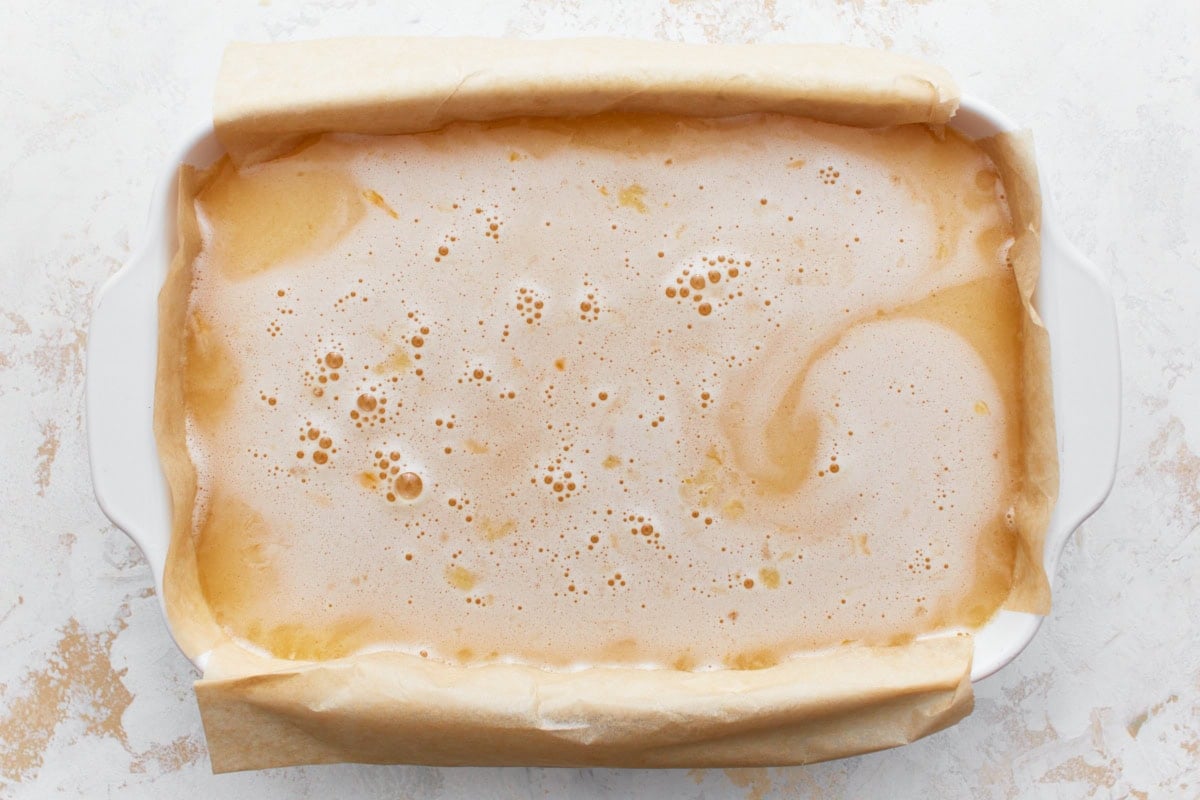 Uncooked lemon filling poured over gluten-free shortbread crust in a parchment paper lined baking dish.
