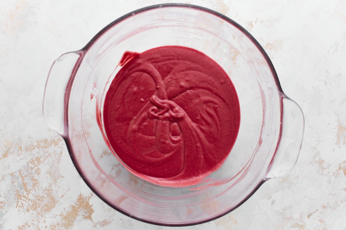A glass mixing bowl containing gluten free red velvet cake batter sitting on a textured white and beige surface.






