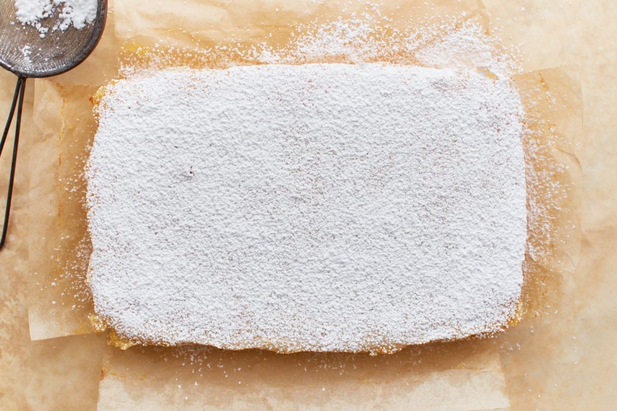 Baked and set gluten-free lemon bars, uncut, laying on parchment paper and dusted with powdered sugar