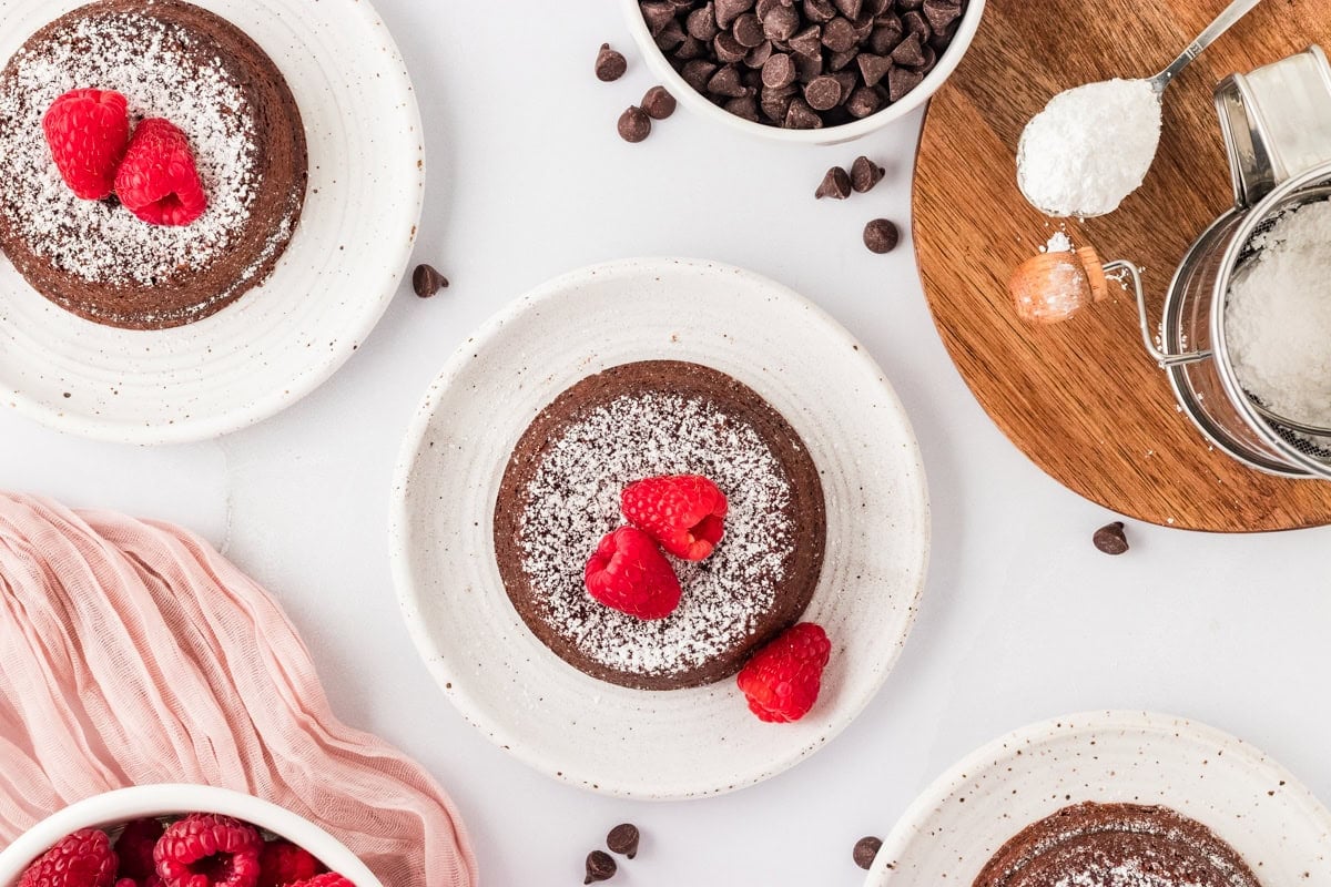 Two gluten-free chocolate lava cakes presented on white plates, topped with powdered sugar and fresh raspberries. Surrounding elements include bowls of raspberries, chocolate chips, and a soft pink fabric, adding visual warmth to the setup.