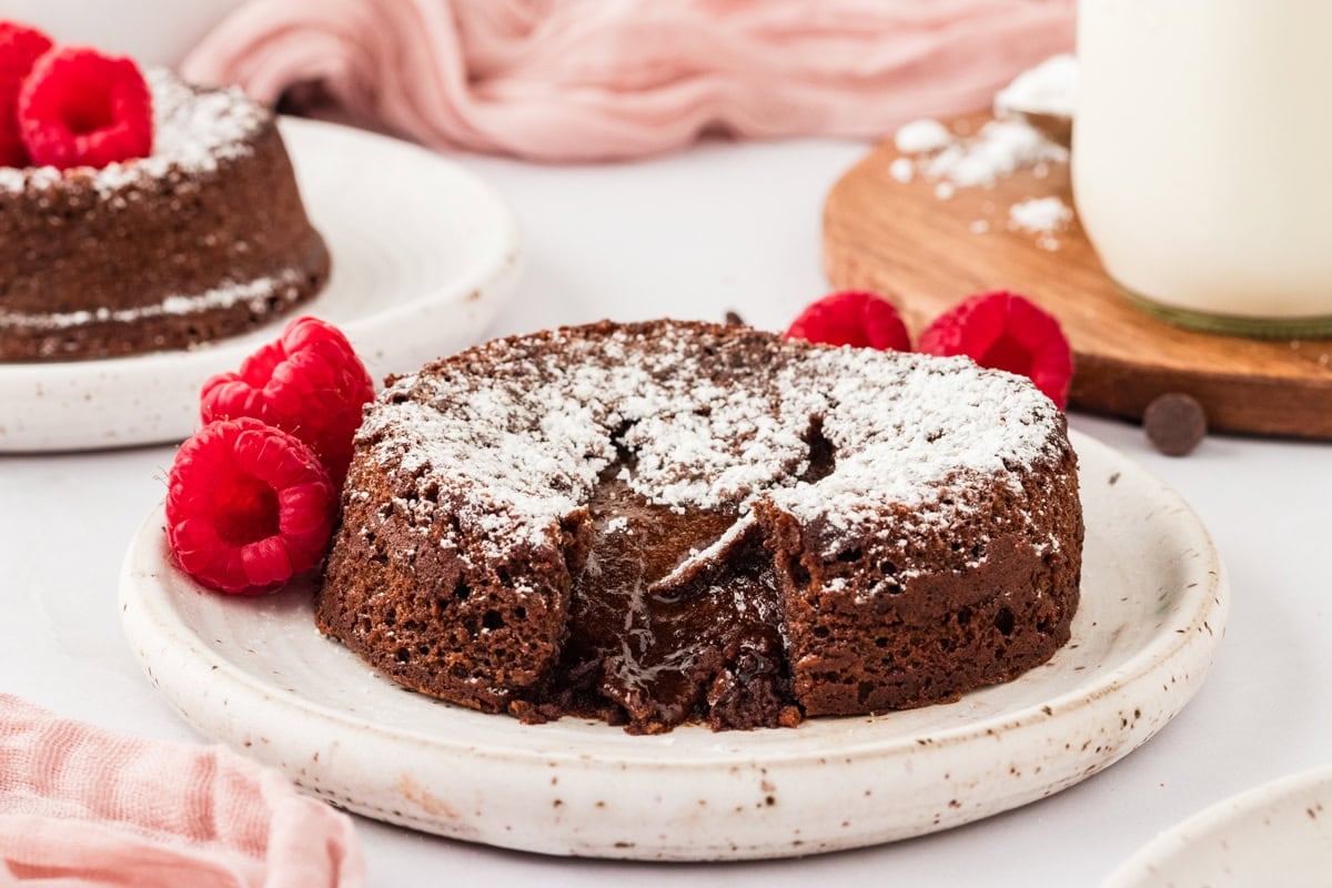 A gluten-free chocolate lava cake on a white plate, dusted with powdered sugar with a gooey chocolate center with raspberries resting on the plate.