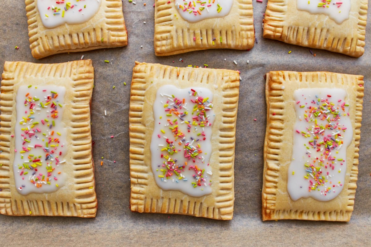 Freshly baked gluten-free pop tart with crimped edges, topped with white icing and colorful sprinkles, placed on parchment paper.