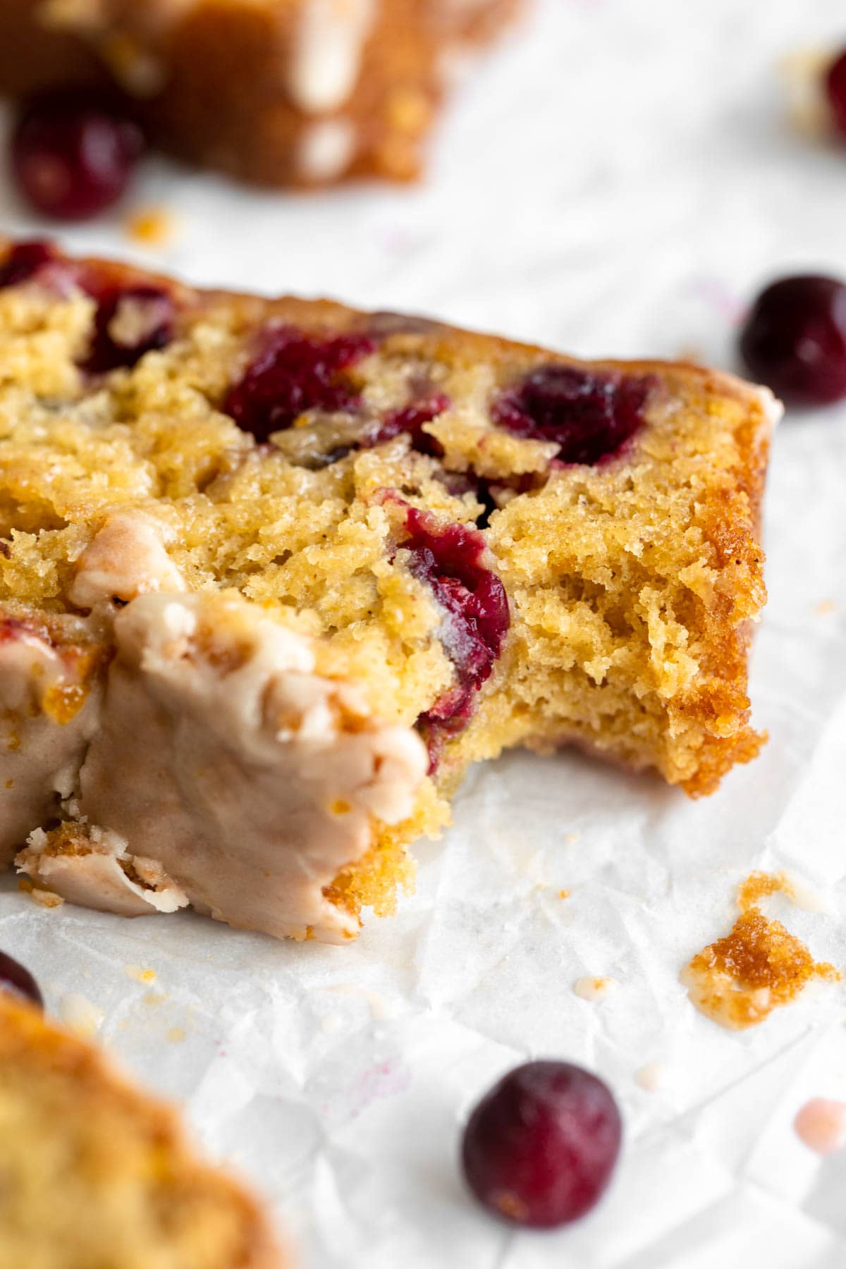 A close-up of a slice of gluten-free cranberry bread with bright red cranberries embedded in a moist, golden crumb. A bite has been taken out, showing its tender texture, and some frosting glaze is visible on the edge