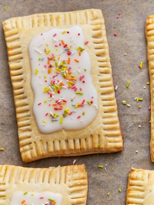 A gluten-free pop tart with a golden crust, topped with white icing and colorful sprinkles, displayed on a parchment-lined surface alongside additional pop tarts.