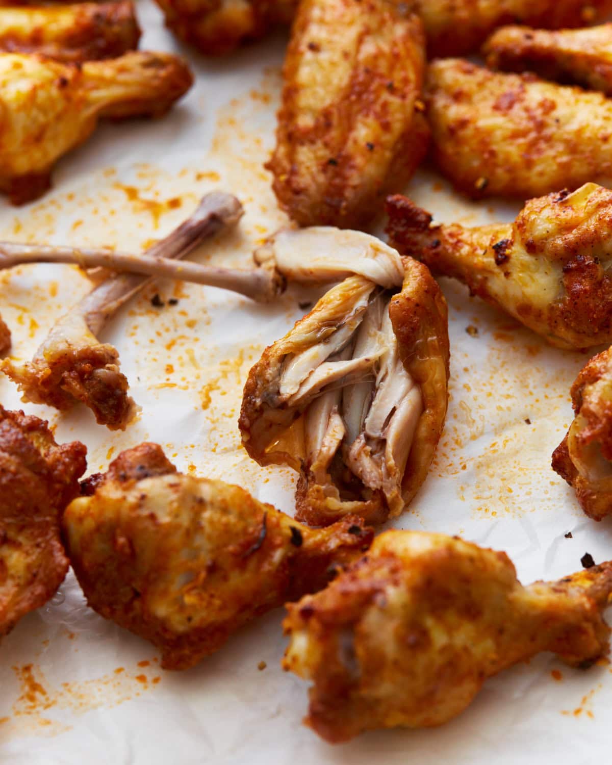 Close-up of crispy, golden-brown  gluten-free chicken wings on parchment paper, with one wing torn open to reveal tender, juicy meat inside. Light seasoning and oil drippings are visible on the surface.