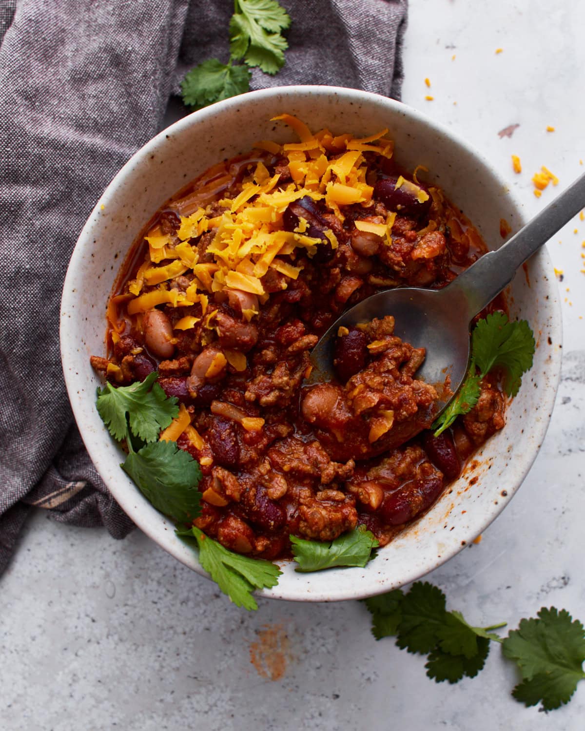 A black spoon digging into a white bowl filled with gluten-free chili, topped with parsley and shredded orange cheese.