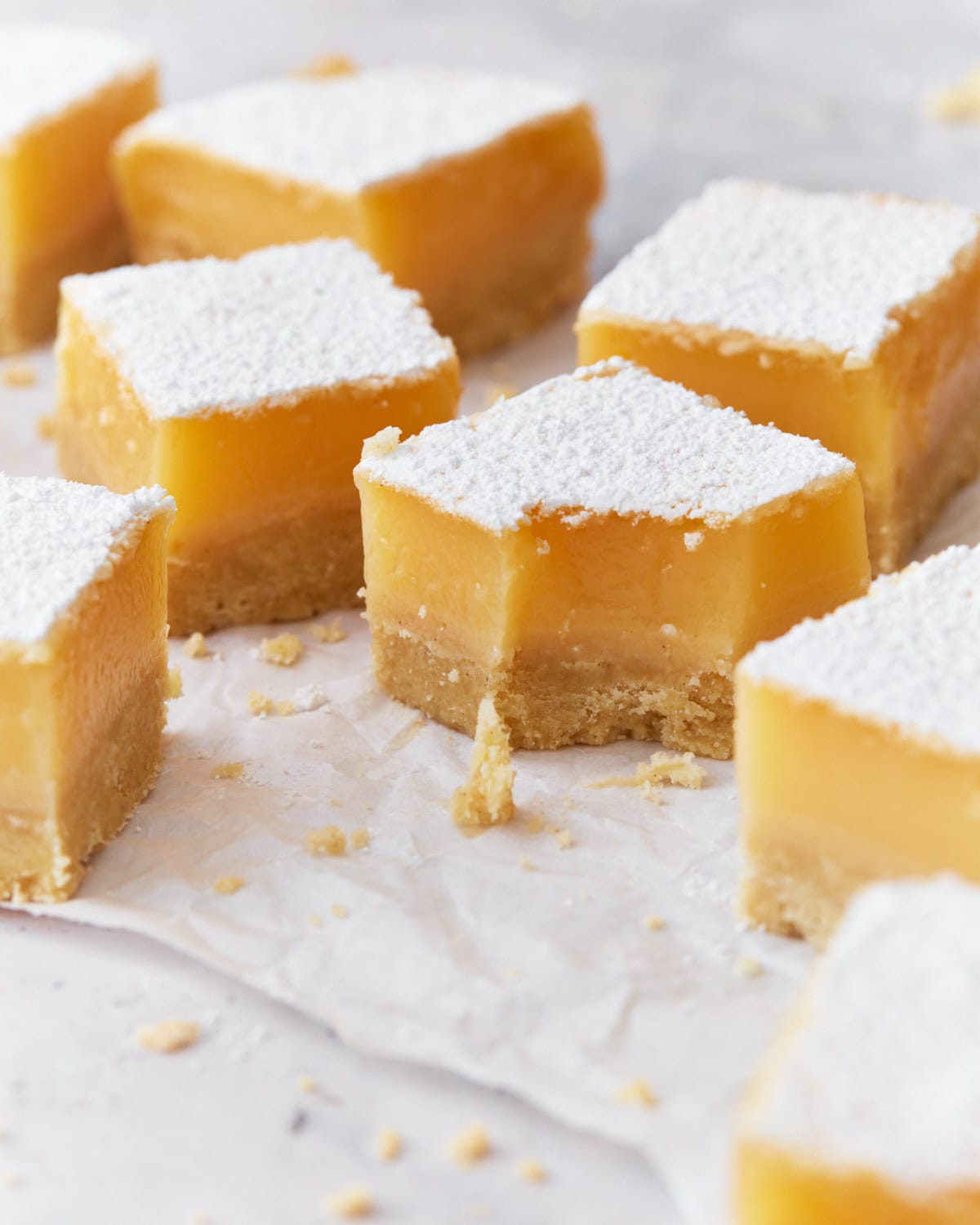 Close-up of gluten-free lemon bars with a thick lemon filling and shortbread crust, dusted with powdered sugar. One bar has a bite taken out, showing the creamy texture, with crumbs scattered on parchment paper