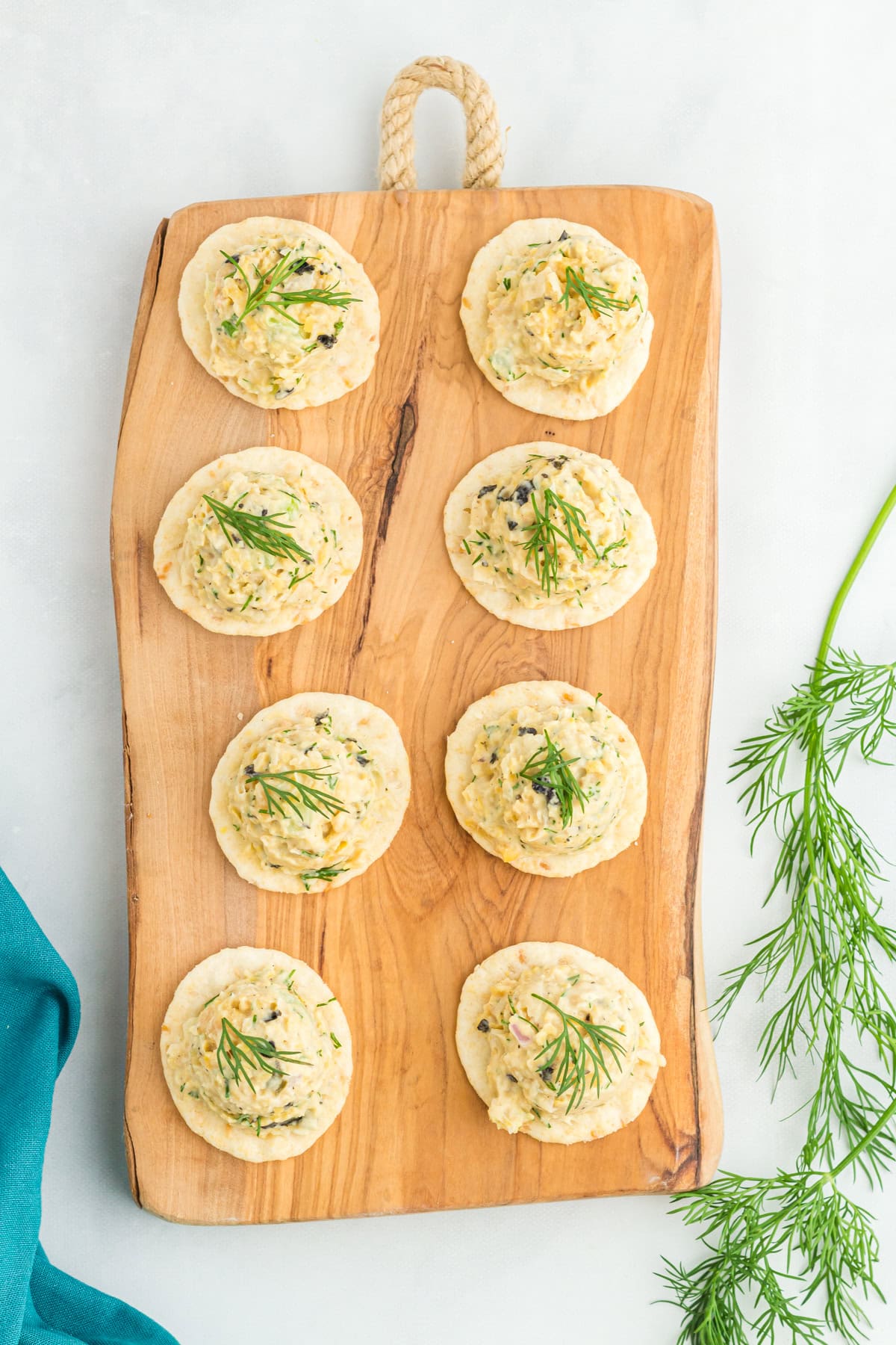 A wooden serving board with eight crackers topped with vegan chickpea tuna salad, garnished with fresh dill. The background includes a sprig of dill and a teal napkin on a white surface