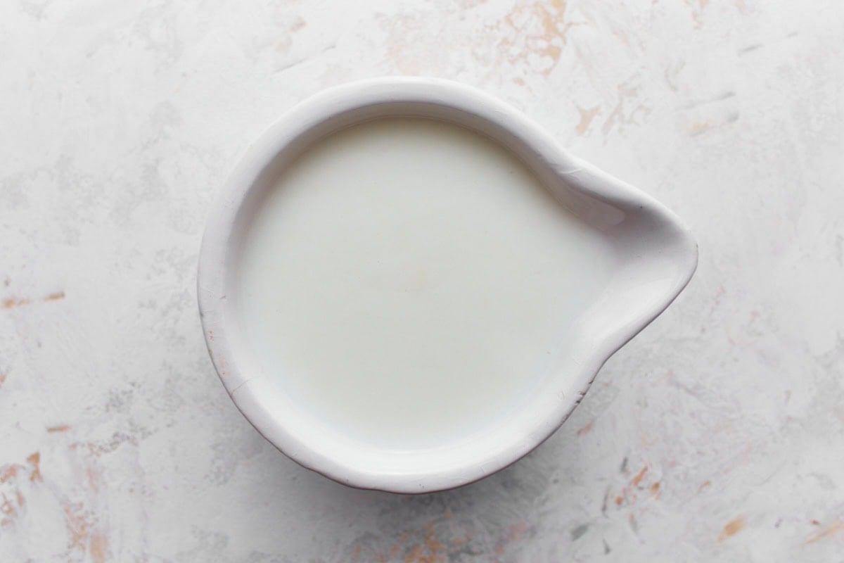 A top-down view of a small white ceramic bowl with a spout, filled with fresh white buttermilk, placed on a textured white and gray surface