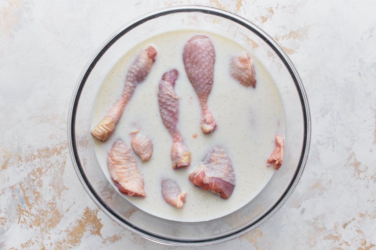 Raw chicken drumsticks and thighs marinating in buttermilk in a large glass bowl
