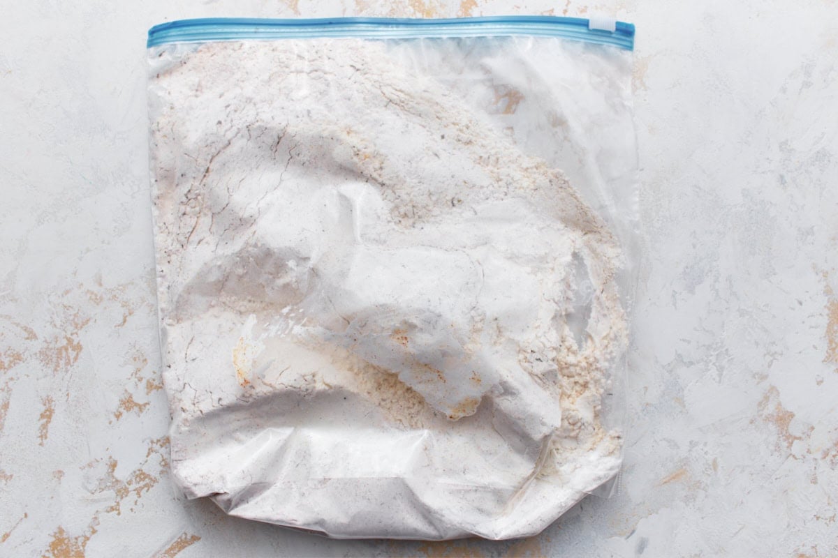 Chicken thighs and drumsticks covered in gluten-free flour, cornstarch, and spice mixture in a large plastic bag laid flat on a light surface.