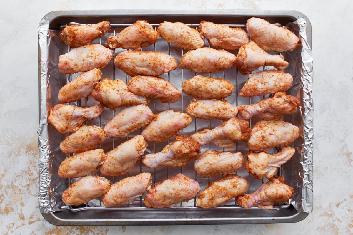 raw chicken wings coated in a dry rub lined up on a wire rack on top of a baking sheet
