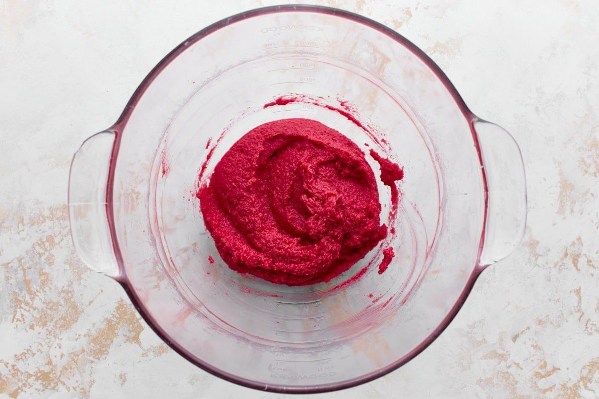 A glass mixing bowl containing vibrant red creamed butter, thick and smooth in texture, sitting on a textured white and beige surface.






