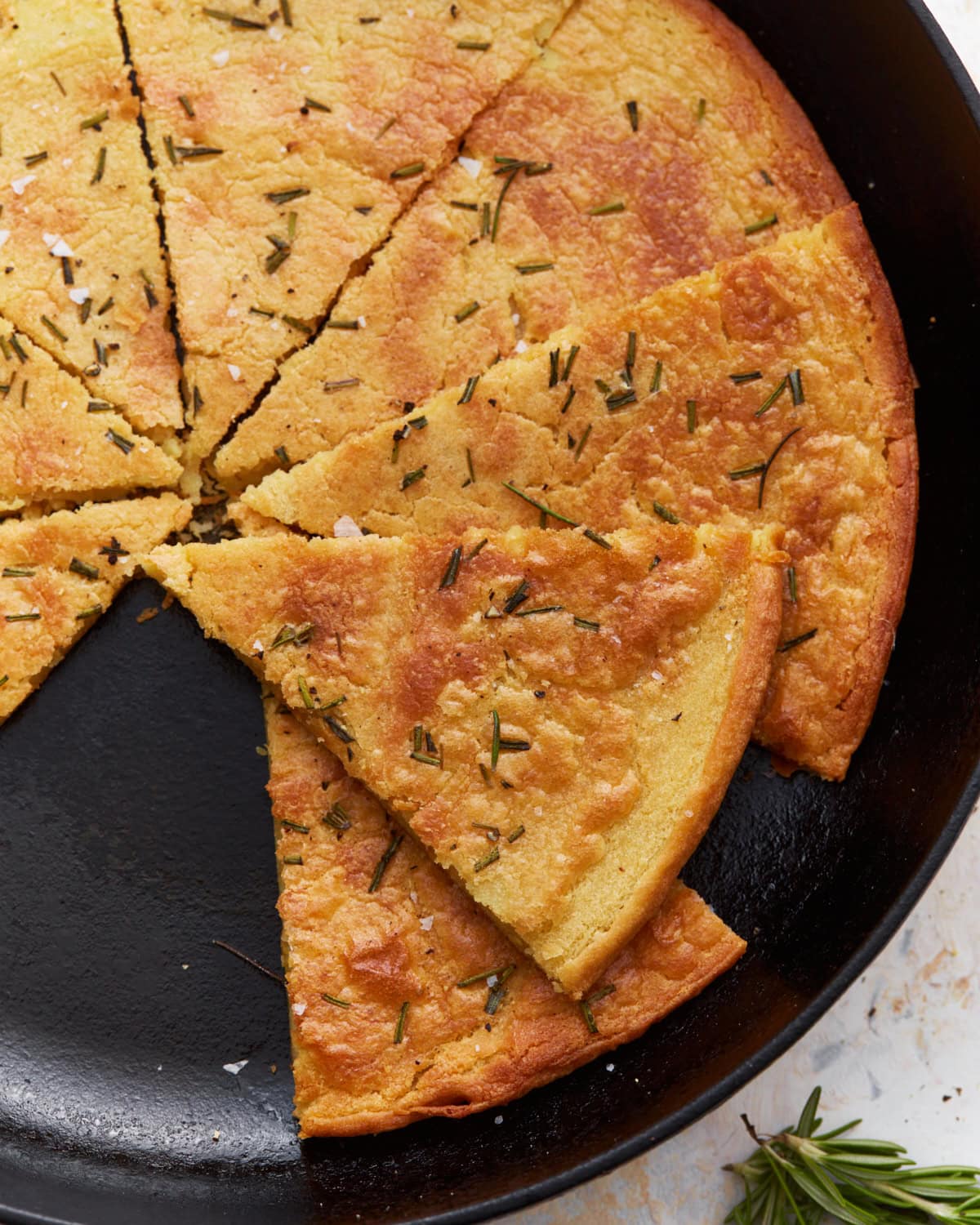 A sliced farinata (chickpea flatbread) in a cast iron skillet, garnished with rosemary and flaky salt.