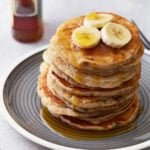 A stack of gluten-free banana pancakes on a gray plate, topped with banana slices and drizzled with maple syrup. A bottle of syrup is blurred in the background.