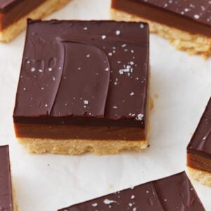 Close-up of a slice of gluten-free millionaire shortbread topped with glossy chocolate, a layer of caramel, and a shortbread base, sprinkled with flaky sea salt.