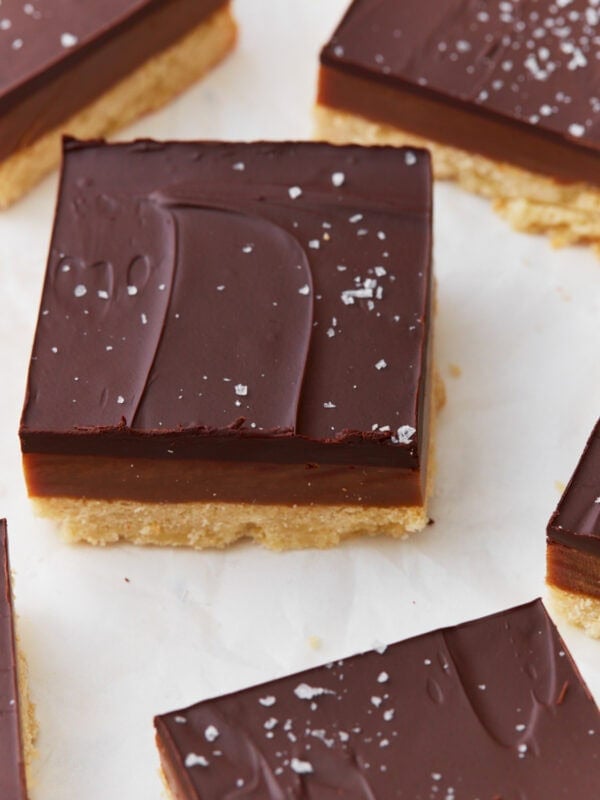 Close-up of a slice of gluten-free millionaire shortbread topped with glossy chocolate, a layer of caramel, and a shortbread base, sprinkled with flaky sea salt.
