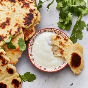 A stack of golden-brown gluten-free naan bread served on a wooden plate, garnished with fresh cilantro leaves, next to a small bowl of creamy yogurt sauce.