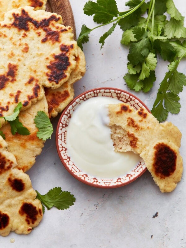 A stack of golden-brown gluten-free naan bread served on a wooden plate, garnished with fresh cilantro leaves, next to a small bowl of creamy yogurt sauce.