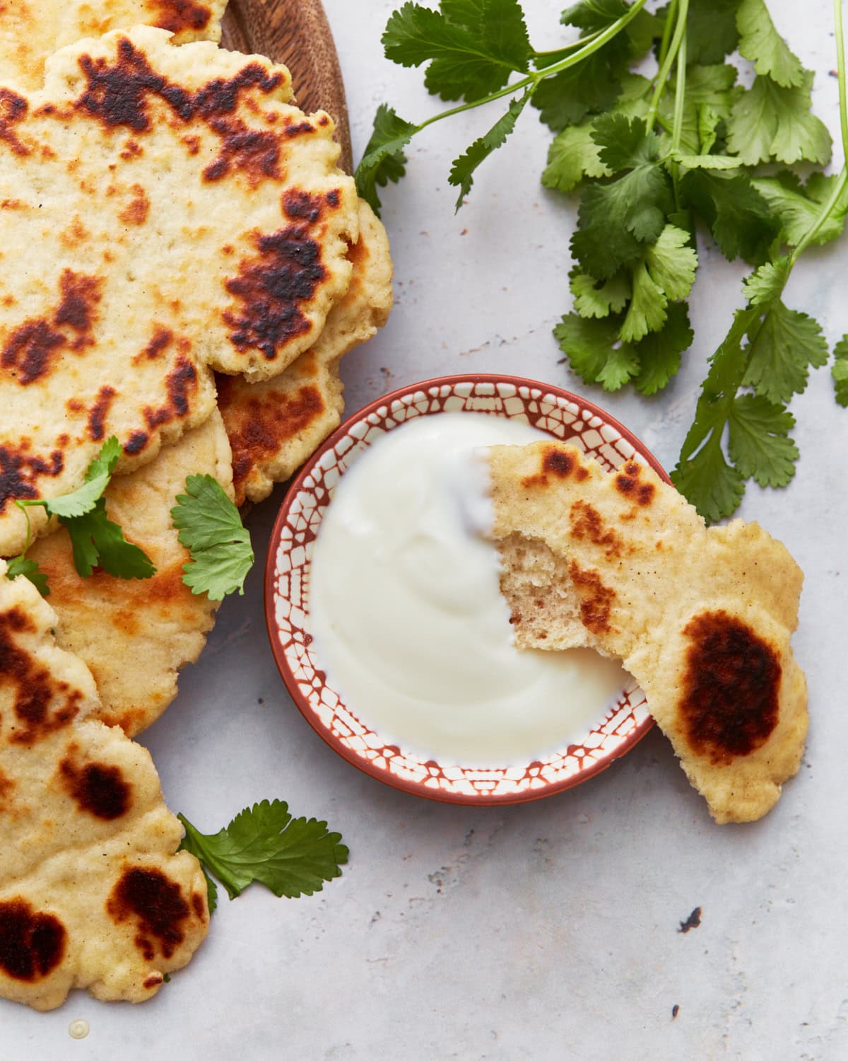 A stack of golden-brown gluten-free naan bread served on a wooden plate, garnished with fresh cilantro leaves, next to a small bowl of creamy yogurt sauce.