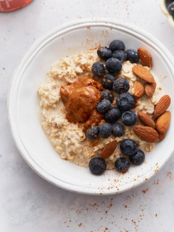 A bowl of creamy gluten-free overnight oats topped with fresh blueberries, whole almonds, and a dollop of almond butter, sprinkled with cinnamon, on a light surface.