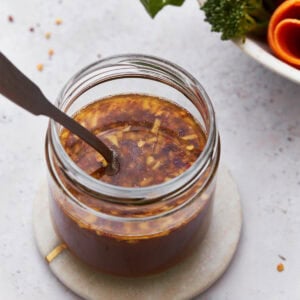 A close-up of a jar filled with gluten-free stir-fry sauce, featuring visible bits of garlic and red chili flakes. The jar sits on a small round coaster, with vibrant fresh broccoli and carrots blurred in the background.