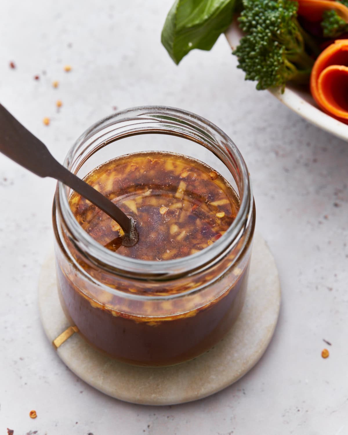 A close-up of a jar filled with gluten-free stir-fry sauce, featuring visible bits of garlic and red chili flakes. The jar sits on a small round coaster, with vibrant fresh broccoli and carrots blurred in the background.