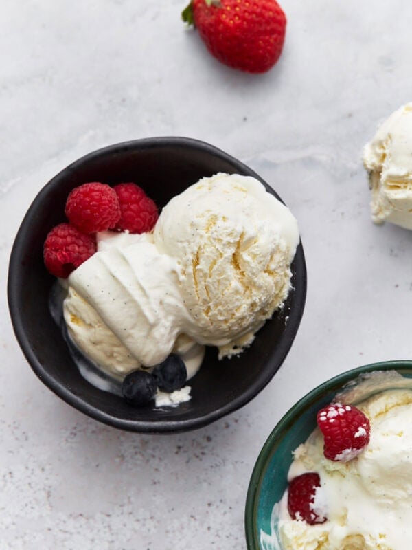 A black bowl filled with scoops of creamy white keto vanilla ice cream, garnished with fresh raspberries and blueberries. A single strawberry sits nearby on the surface.