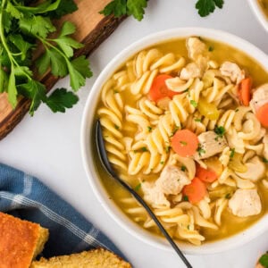 A close-up of a bowl of gluten-free chicken noodle soup featuring spiral noodles, chunks of chicken, carrots, celery, and a clear broth, garnished with fresh parsley, with cornbread and parsley in the background.