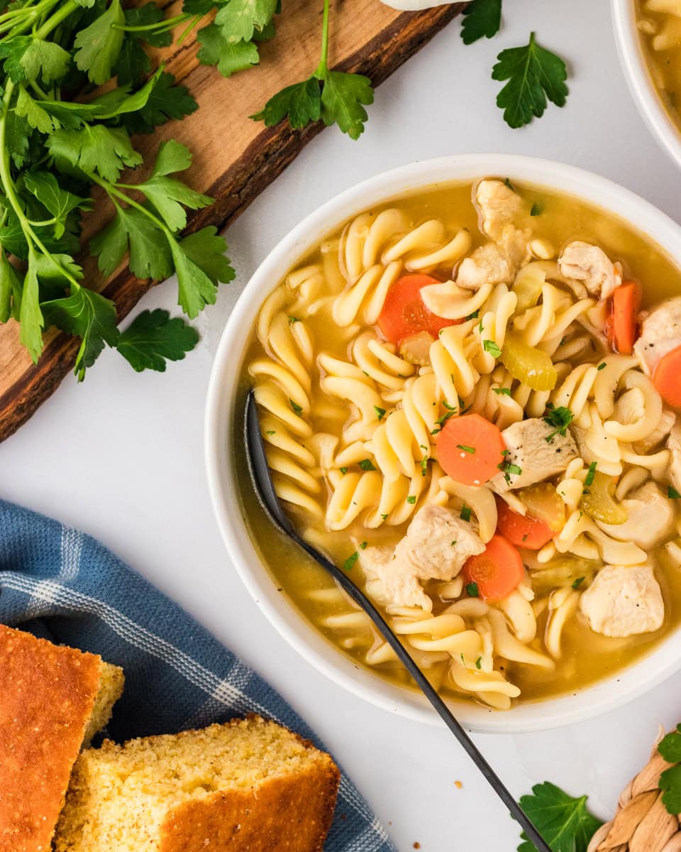 A close-up of a bowl of gluten-free chicken noodle soup featuring spiral noodles, chunks of chicken, carrots, celery, and a clear broth, garnished with fresh parsley, with cornbread and parsley in the background.