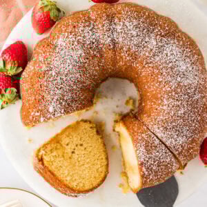 A Gluten-Free Kentucky Butter Pound Cake on a round white platter, topped with powdered sugar and garnished with fresh strawberries. A slice has been cut and partially removed, showcasing the cake's golden interior.