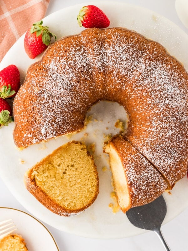 A Gluten-Free Kentucky Butter Pound Cake on a round white platter, topped with powdered sugar and garnished with fresh strawberries. A slice has been cut and partially removed, showcasing the cake's golden interior.